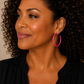A close-up of a woman with curly dark hair wearing a bright pink hoop earring, dressed in a black top against a neutral beige background.