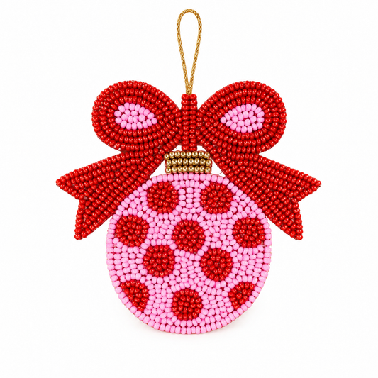 A beaded Christmas ornament shaped like a round bauble with a large red bow on top, hanging from a gold string. The ornament is made of pink and red seed beads with red polka dots on a pink background.