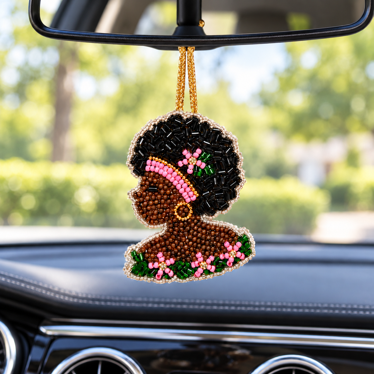 Black Girl Bloom Energy beaded charm with an Afro, pink headband, and pink-and-green flowers hanging from a car’s rearview mirror by a gold cord.