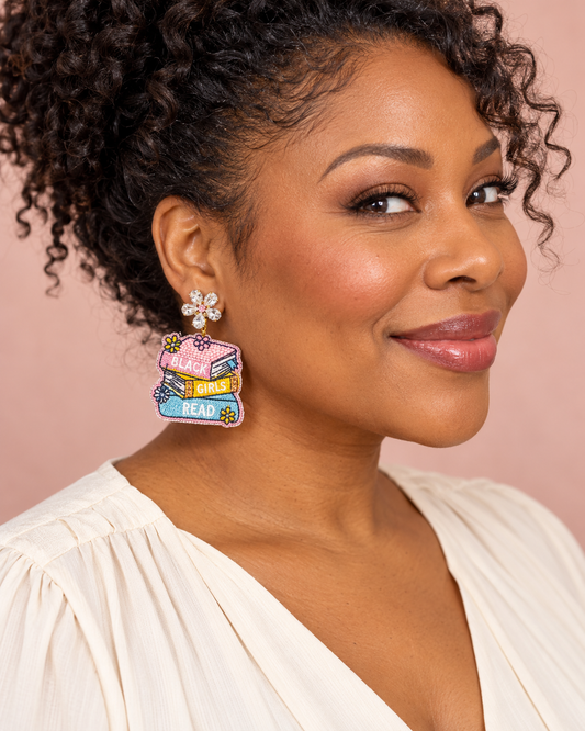 Woman wearing “Black Girls Read” book-stack earrings with crystal flower tops against a blush background.