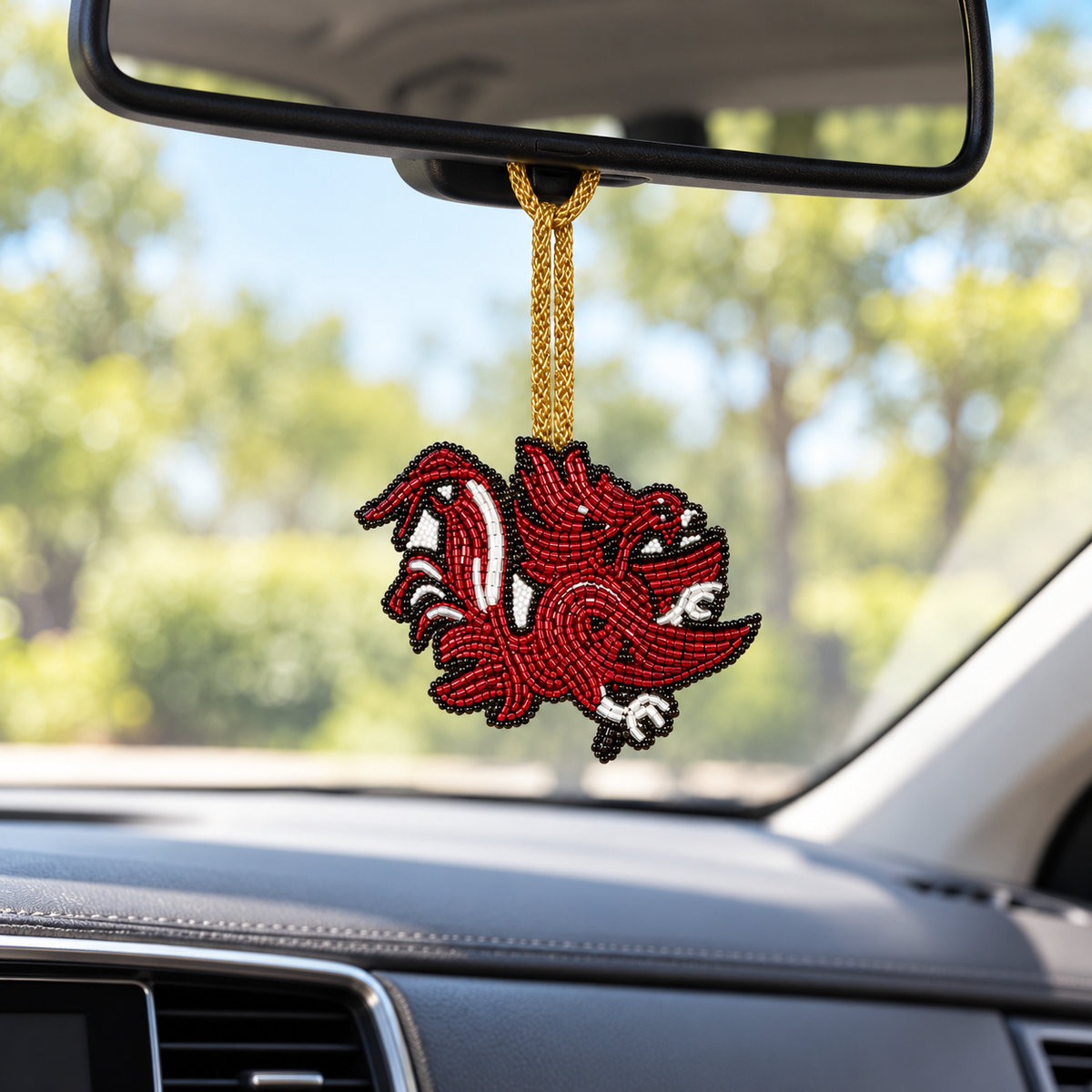 Beaded Gamecock charm in garnet, black, and silver hanging from a car’s rearview mirror by a gold cord.