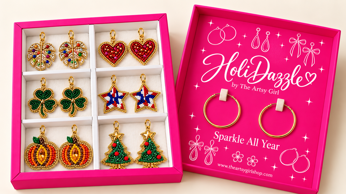 Open hot pink HoliDazzle box with six pairs of beaded holiday charms and one pair of gold hoops beside the divided tray.