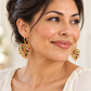 Woman in an ivory blouse wearing gold hoops with openwork gold beaded leaf charms accented with colorful gems.