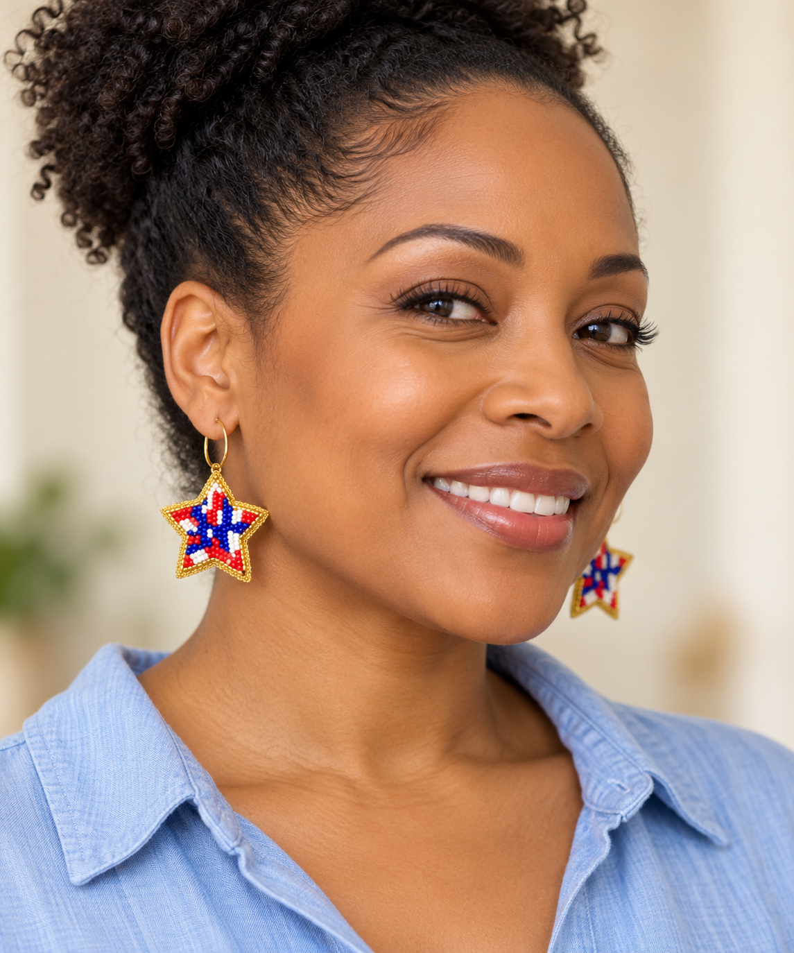 Woman in a light blue shirt wearing gold hoops with red, white, and blue beaded star charms edged in gold.
