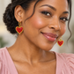 Woman in a blush blouse wearing gold hoops with red and pink beaded heart charms edged in gold.