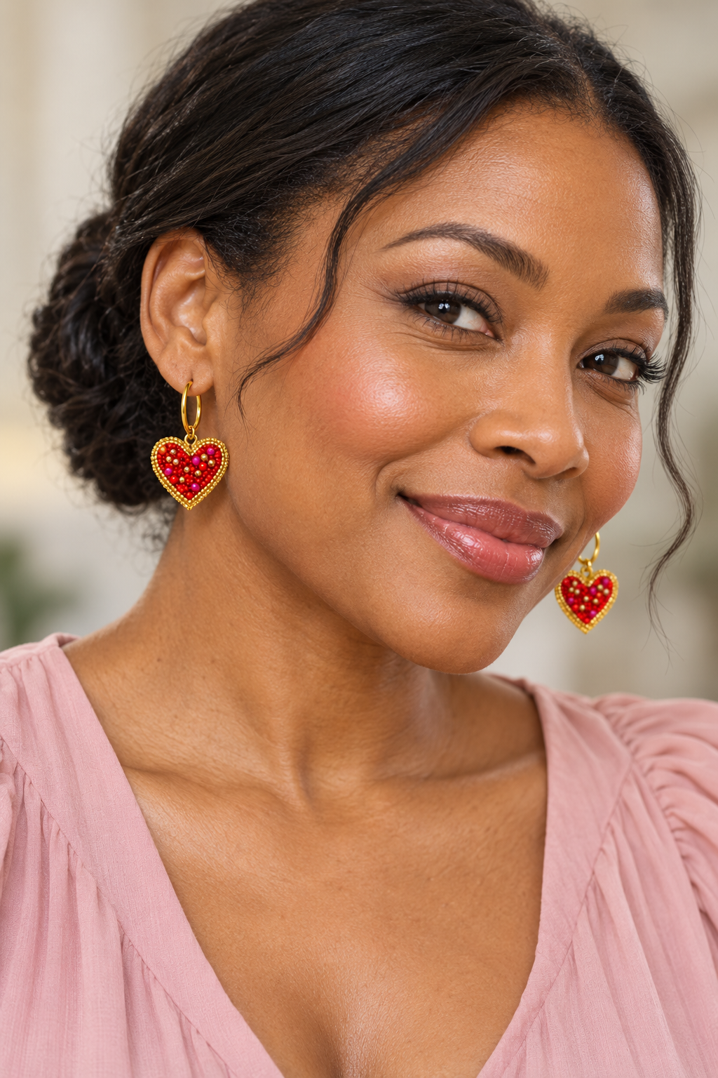 Woman in a blush blouse wearing gold hoops with red and pink beaded heart charms edged in gold.