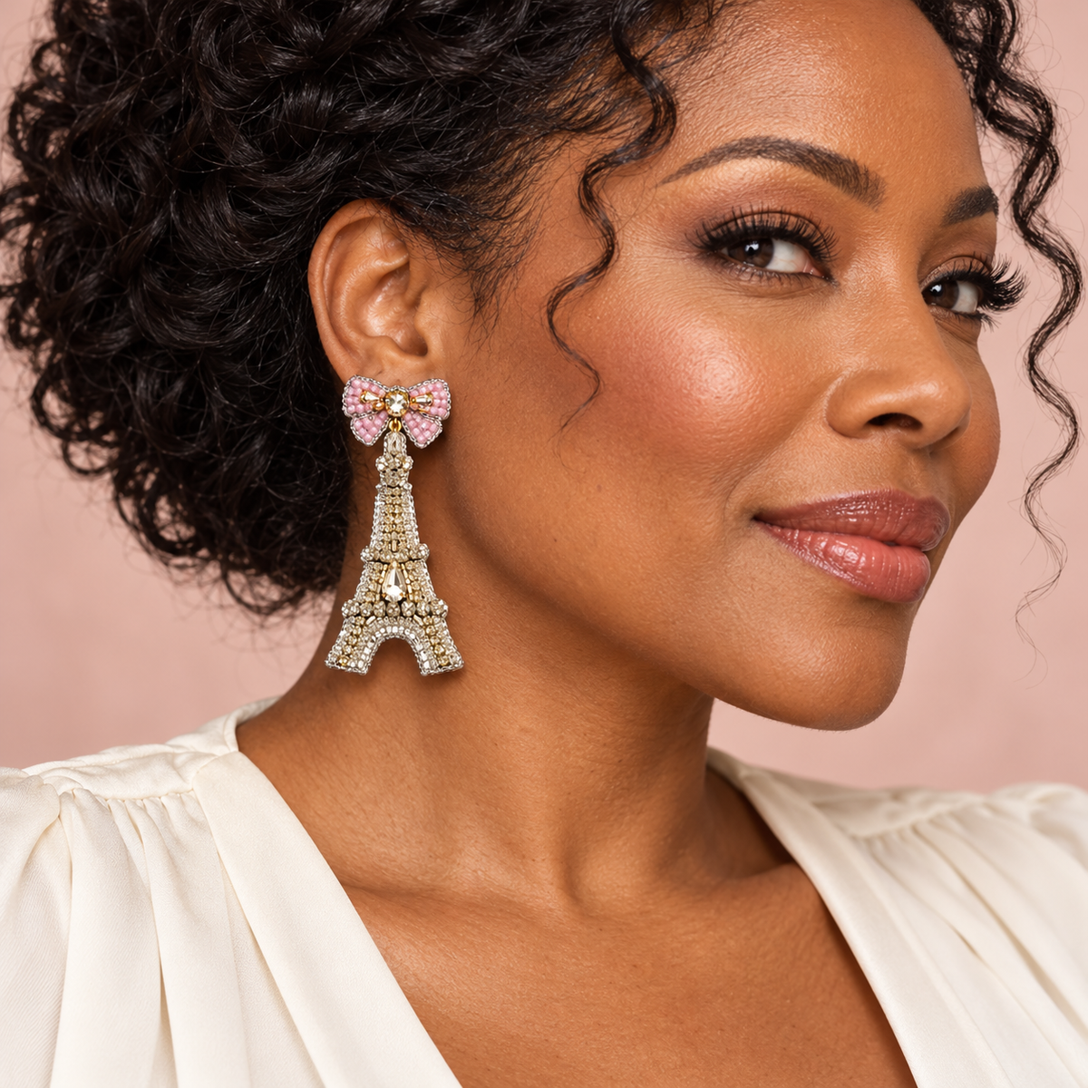 Woman wearing a crystal Eiffel Tower earring with a pink beaded bow stud against a blush background.