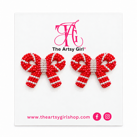 Red and white beaded bow stud earrings on The Artsy Girl display card against a white background.