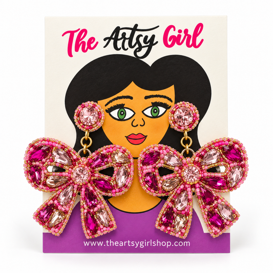Pink beaded bow stud earrings with metallic accents on The Artsy Girl display card against a white background.