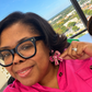 A close-up selfie of a woman wearing a bright pink button-up shirt and black-rimmed glasses, holding up a pink rhinestone bow earring. A cityscape is visible in the background through a large window.