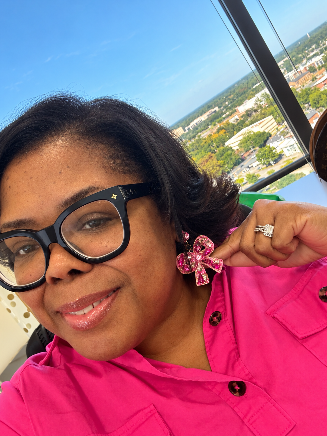 A close-up selfie of a woman wearing a bright pink button-up shirt and black-rimmed glasses, holding up a pink rhinestone bow earring. A cityscape is visible in the background through a large window.