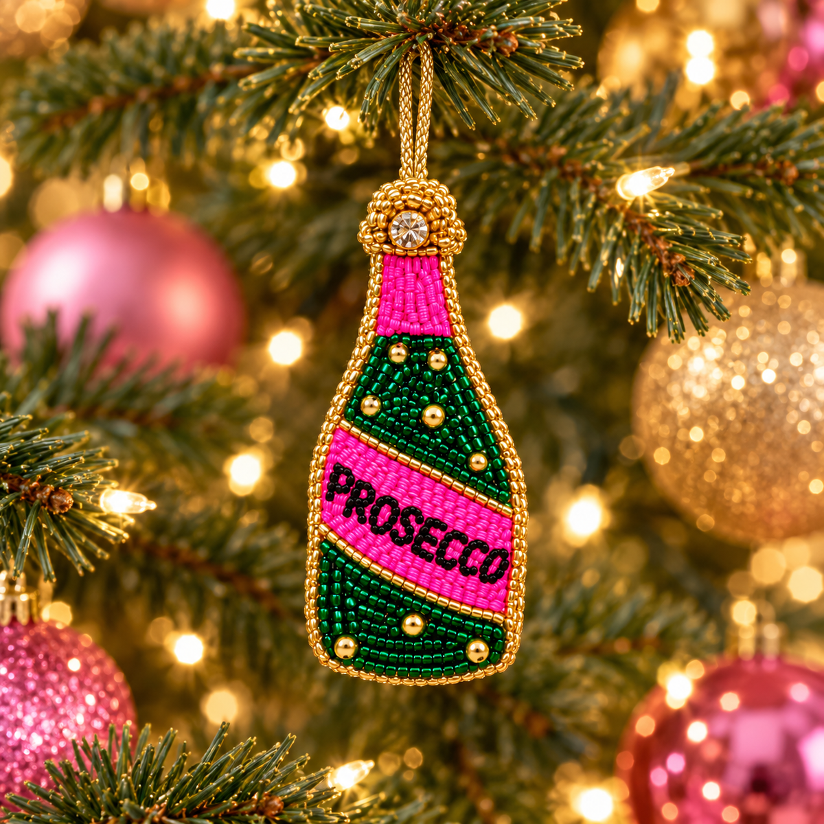 Beaded Prosecco bottle charm hanging on a Christmas tree with pink and gold ornaments and warm lights.