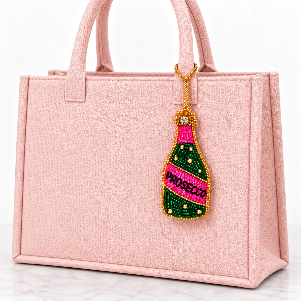 Pink and green beaded Prosecco bottle charm hanging from a light pink tote by a gold cord.