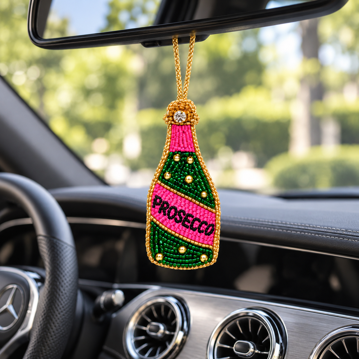 Pink and green beaded Prosecco bottle charm hanging from a rearview mirror inside a Mercedes.