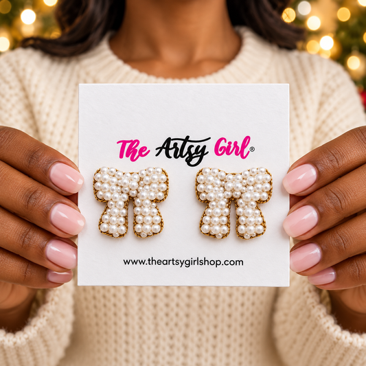 Woman holding The Artsy Girl card with white pearl bow earrings against softly blurred holiday lights.